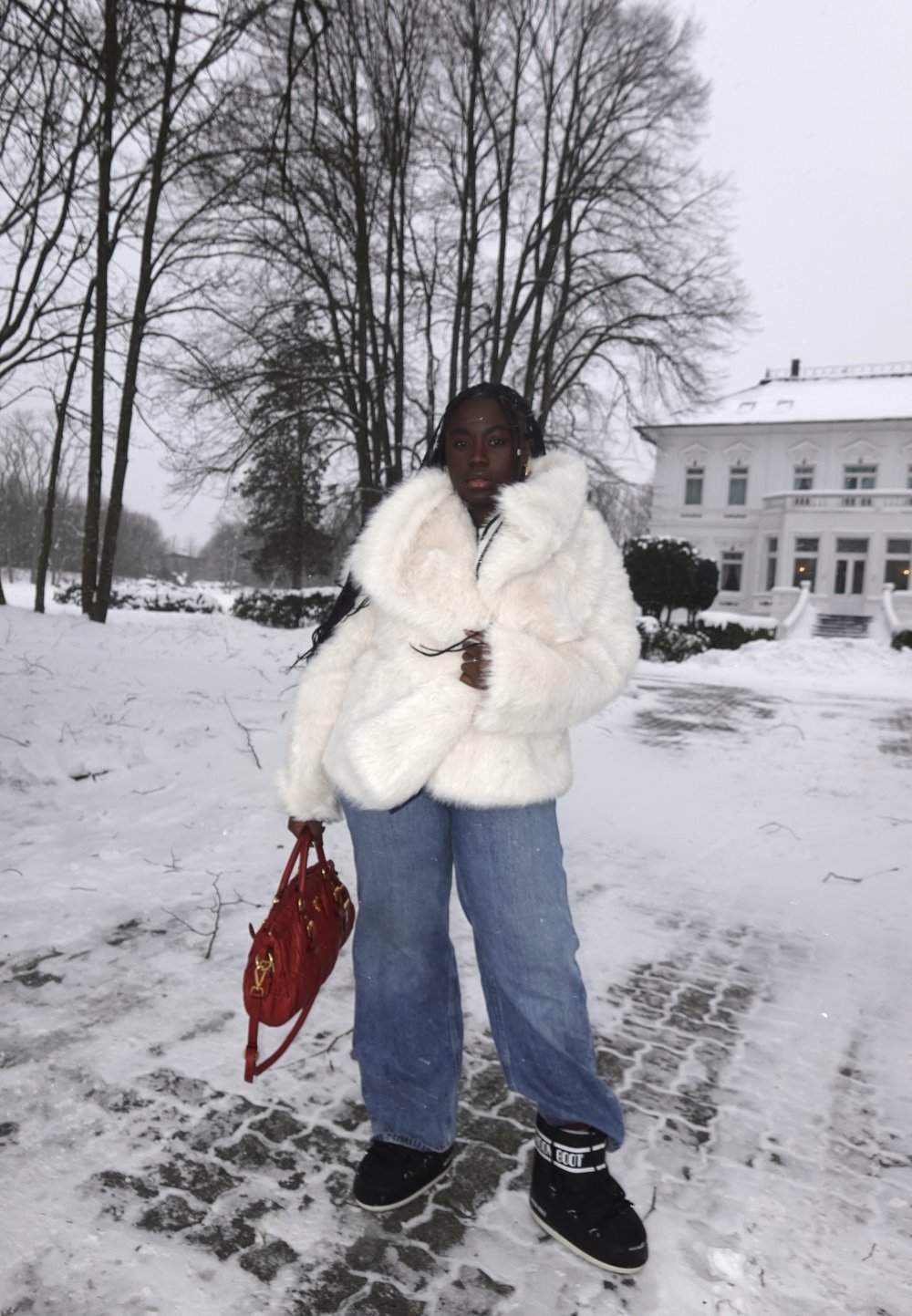 Femme debout sur un chemin enneigé portant un manteau en fourrure blanc, un jean bleu, des bottes noires, tenant un sac à main rouge, avec des arbres sans feuilles et un bâtiment blanc en arrière-plan.