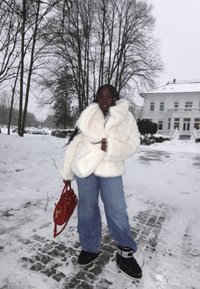 Femme debout sur un chemin enneigé portant un manteau en fourrure blanc, un jean bleu, des bottes noires, tenant un sac à main rouge, avec des arbres sans feuilles et un bâtiment blanc en arrière-plan.