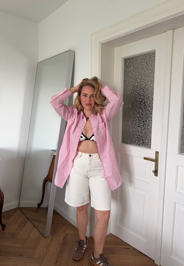 Frau steht drinnen mit den Händen im Haar, trägt ein pinkes Hemd über einem schwarz-weißen Bikini-Top, weiße Shorts und braune Turnschuhe.