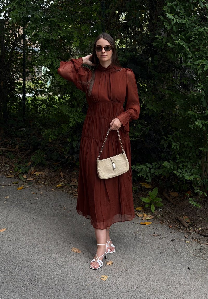 Femme portant une longue robe couleur rouille, des sandales blanches à brides et des lunettes de soleil foncées, tenant un sac à main beige, debout sur un chemin près d'un feuillage vert.