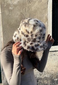 Eimerhut aus Kunstpelz in Beige mit schwarzen und braunen Leopardenflecken. Weiche Textur, abgerundete Oberseite und breite Krempe für Schutz.