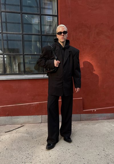 Blazer negro de gran tamaño sobre un hoodie negro, combinado con pantalones de pierna ancha y zapatos negros brillantes. Las gafas de sol y un bolso negro completan el look.