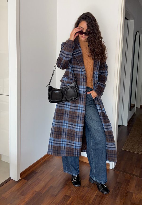Une femme ajuste ses lunettes de soleil tout en posant à l’intérieur avec un long manteau bleu à carreaux, un jean et un sac noir porté à l’épaule.