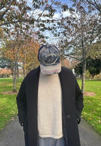 Fekete kabát világos bézs pulóver fölött, szürke melegítőnadrág és kopott kék sapka logóval, őszi falevelekkel teli fák előtt.