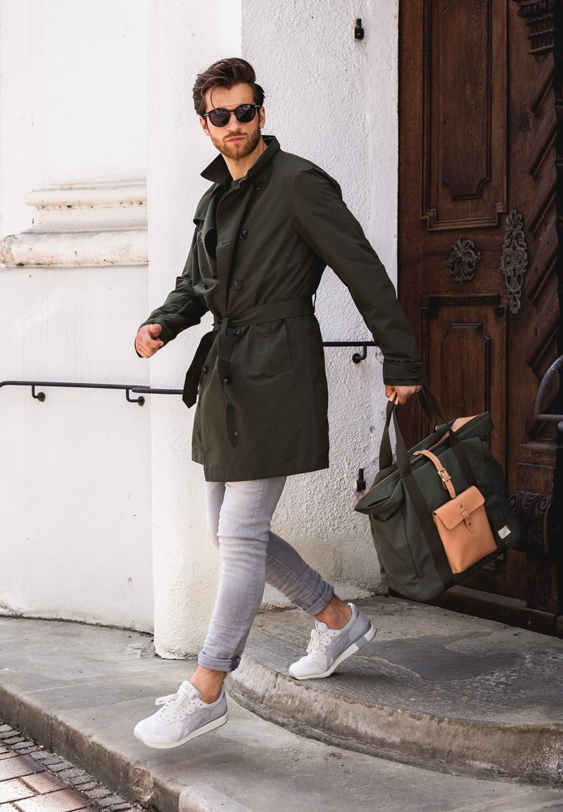 Olijfgroen trenchcoat met zwarte knopen, lichtgrijze jeans, witte sneakers en een twee-kleurige tas met donkergroene en tan accenten.