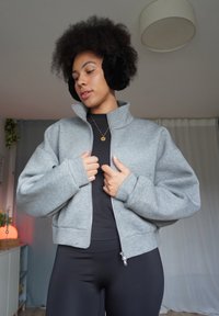 Grey zip-up sweatshirt with high collar and loose sleeves, worn over a black top, paired with black leggings. Soft fabric with smooth texture.