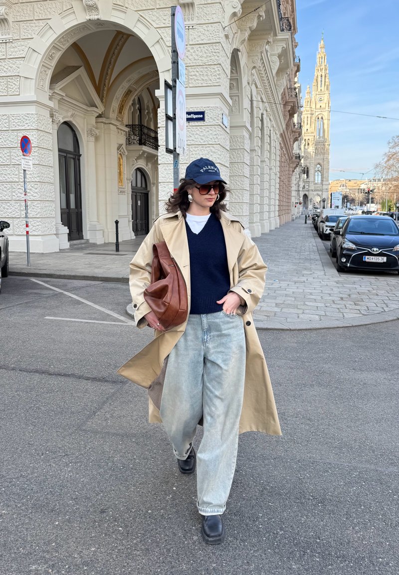 Donna con trench beige, jeans oversize, occhiali da sole e cappellino che cammina per strada in città vicino a un edificio storico e auto parcheggiate sotto un cielo sereno.