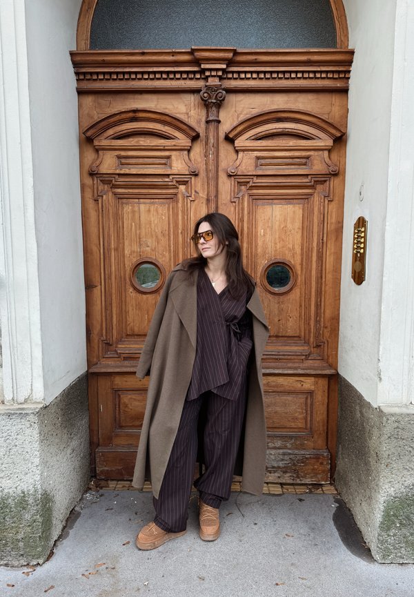 Brown wool coat over a dark striped wrap top and wide-leg trousers. Features wooden double doors with circular glass panels. Brown platform shoes.