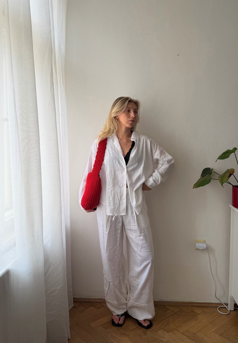 Femme en chemise et pantalon blancs se tient près d'une fenêtre, portant des sandales noires et un sac bandoulière rouge tricoté, avec une plante dans le coin.