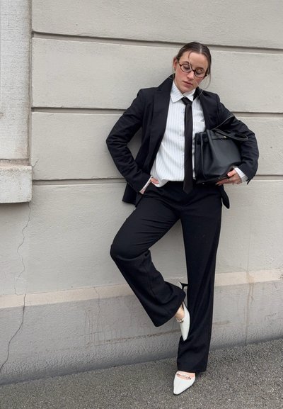 Mujer con traje negro, camisa blanca y corbata, apoyada en la pared, sosteniendo un bolso negro, con gafas y zapatos blancos puntiagudos con tacón.