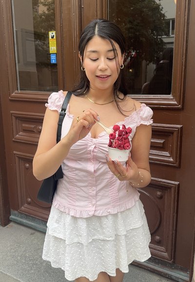 Copa de helado de frambuesa decorada con trozos de frambuesa, sostenida por una persona con una blusa rosa claro y una falda blanca con volantes, frente a una puerta marrón.