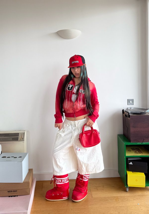 Jeune personne posant à l'intérieur vêtue d'une casquette rouge Adidas, d'une veste rouge, d'un pantalon blanc large, de bottes de neige rouges Moon Boot, et tenant un sac à main rouge.