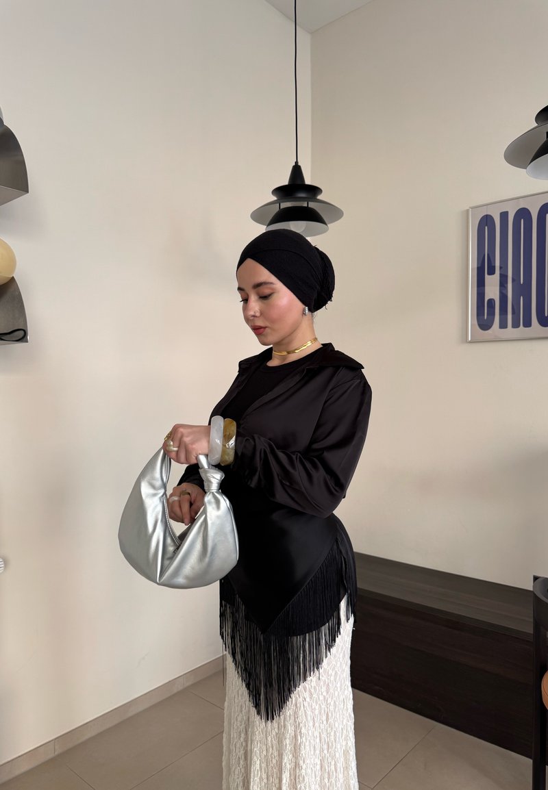 Top en satin noir avec des franges, associé à une jupe texturée blanche. Présente un sac hobo métallique argenté et des accessoires ronds.
