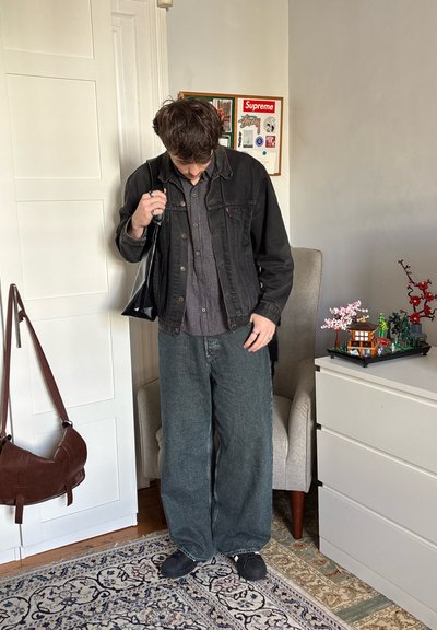 Chaqueta de mezclilla negra sobre una camisa de cuadros gris, combinada con jeans oscuros verdes holgados y zapatos negros, sosteniendo un bolso negro. Fondo neutro.