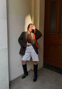 Sort blazer over en brun cropped sweater, hvide shorts, sorte knæhøje støvler med strikdetaljer og en orange håndtaske, i et godt oplyst hjørne.