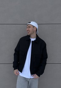 Un homme souriant regardant vers la droite, portant une casquette blanche à l'envers, une veste noire, une chemise blanche et un jean gris clair, debout contre un mur gris.