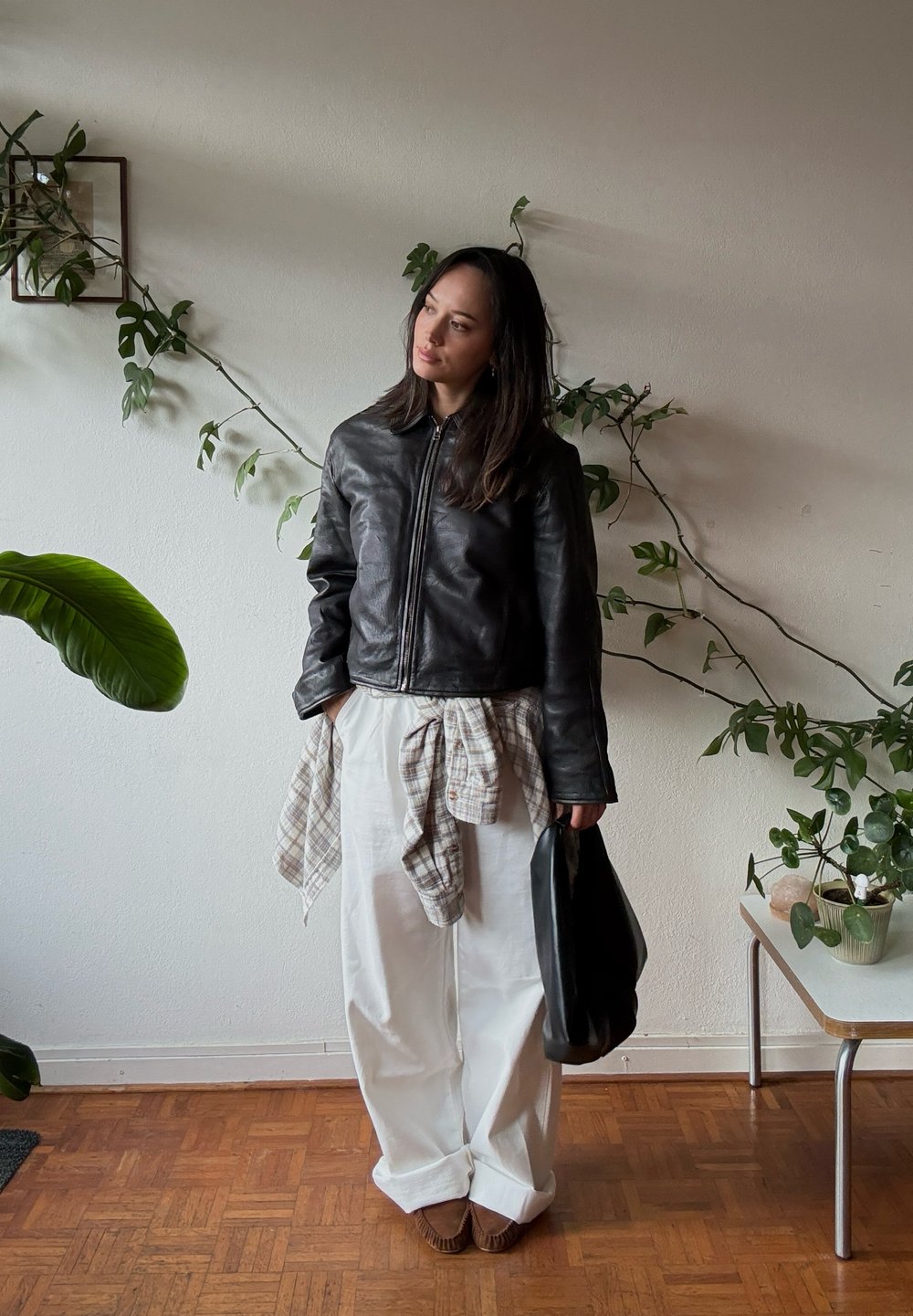 Giacca di pelle nera con zip frontale, abbinata a pantaloni bianchi larghi, una camicia a quadri legata in vita e una borsa nera.