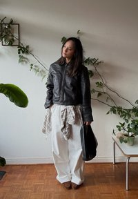 Giacca di pelle nera con zip frontale, abbinata a pantaloni bianchi larghi, una camicia a quadri legata in vita e una borsa nera.