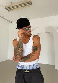 Camisola de alças, óculos de sol cinzentos, boné preto e calças de moletom com faixa azul; braços cobertos por tatuagens detalhadas; fundo neutro.