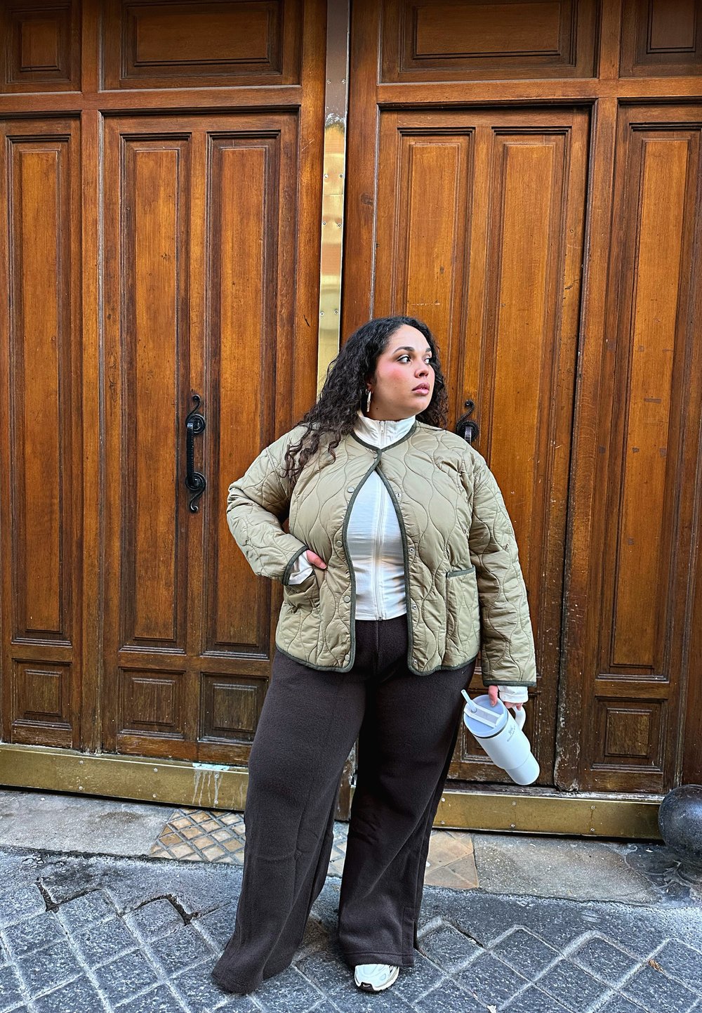 Khakifarbenes gesteppte Jacket mit zwei Taschen, weißes Hochkragenhemd, braune weite Hose und weiße Sneaker. Im Hintergrund sind Holztüren zu sehen.