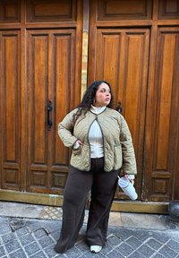 Khakifarbenes gesteppte Jacket mit zwei Taschen, weißes Hochkragenhemd, braune weite Hose und weiße Sneaker. Im Hintergrund sind Holztüren zu sehen.