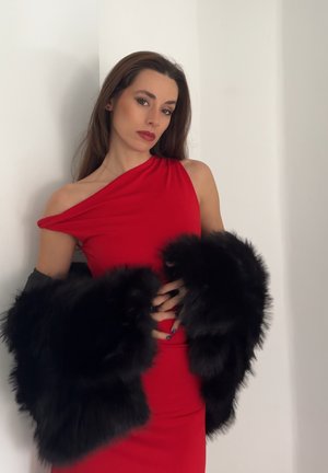 Mujer con cabello largo y castaño, vestida con un vestido rojo de un solo hombro, sosteniendo una estola de piel negra contra un fondo blanco liso.