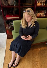 Une personne assise sur un canapé vert, portant une chemise longue noire et une jupe en denim bleue, utilisant un smartphone, avec des lunettes de soleil sur la tête.