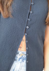 Temno mornarsko modra teksturirana bluza z gumbom spredaj in stranskim razporkom, s sivimi gumbi in lahkim materialom, v kombinaciji z modrimi jeans kratkimi hlačami.