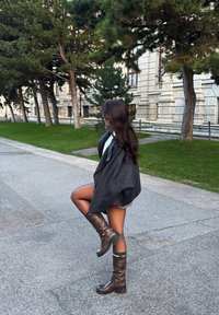 Stivali in pelle marroni fino al ginocchio con fibbie, cappotto grigio oversize, dolcevita bianca e capelli scuri, ambientati in un parco con edifici sullo sfondo.