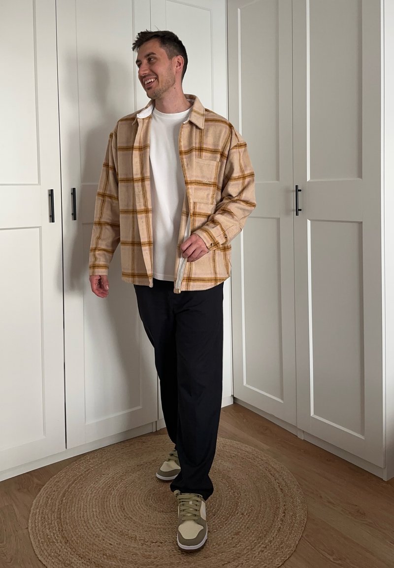 Giacca in flanella a quadri marrone su una camicia bianca, abbinata a pantaloni neri e sneakers beige. Tessuto testurizzato con bottoni e tasche visibili.