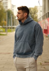 Modrá bavlněná mikina s kapucí a přední kapsou, nošená s béžovými tepláky. Tkanina vykazuje mírně vybledlou strukturu.