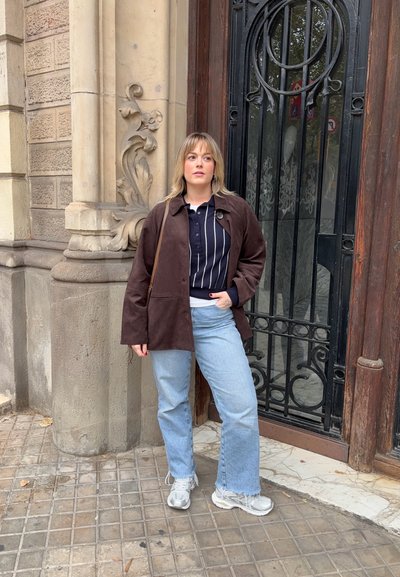 Chaqueta de ante marrón sobre un polo a rayas blancas y negras, combinado con unos vaqueros de mezclilla azul claro y zapatillas plateadas. De pie frente a un edificio decorativo.