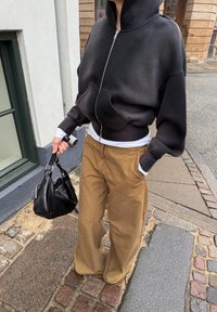 Άτομο με μαύρο φούτερ με φερμουάρ, λευκό πουκάμισο, καφέ παντελόνι φαρδύ στο κάτω μέρος, κρατώντας μαύρη τσάντα χειρός, στέκεται σε πλακόστρωτο δρόμο.