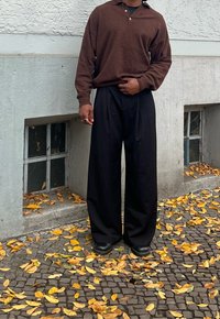 Osoba ubrana w brązową koszulę z długim rękawem, czarne spodnie o szerokich nogawkach i czarne buty stoi na chodniku z kostki brukowej pokrytym liśćmi.