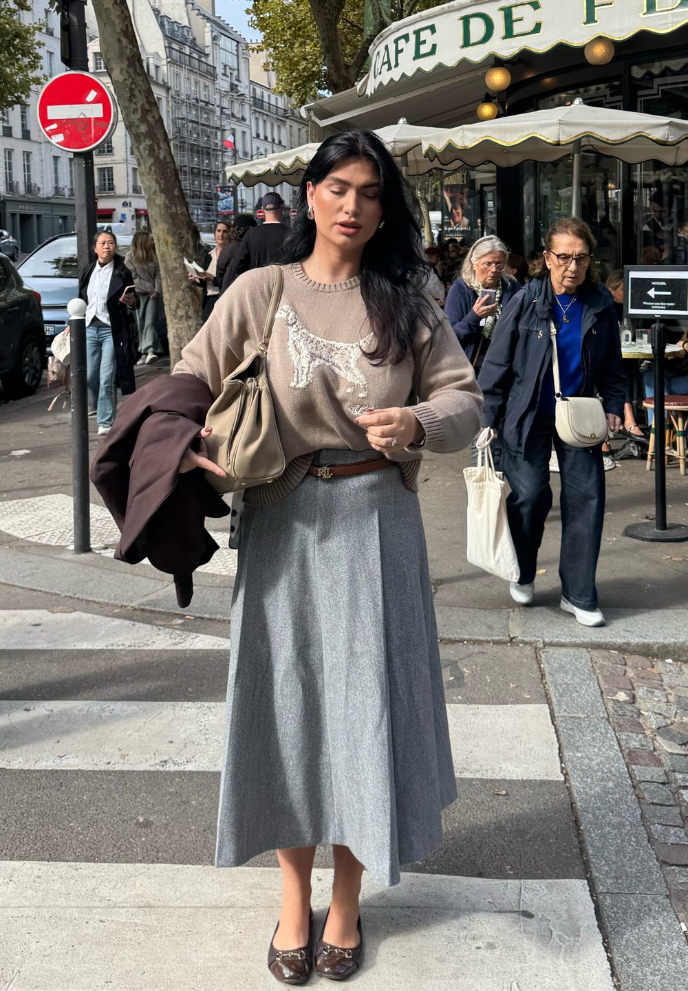 Hellbrauner Pullover mit Hundef design, grauer Faltenrock und braunen Schuhen. Hält eine beiger Tasche und hat einen dunklen Mantel über dem Arm.