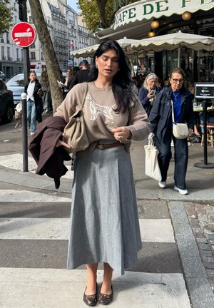 Jersey marrón claro con un diseño de perro, falda plisada gris y zapatos marrones. Sosteniendo un bolso beige, con un abrigo oscuro colgado de su brazo.