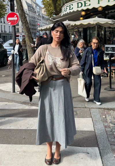 Jersey marrón claro con un diseño de perro, falda plisada gris y zapatos marrones. Sosteniendo un bolso beige, con un abrigo oscuro colgado de su brazo.