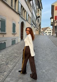 Pantaloni sportivi marroni con strisce gialle, abbinati a una maglia bianca a maniche lunghe. L'ambientazione presenta una strada lastricata e edifici storici.