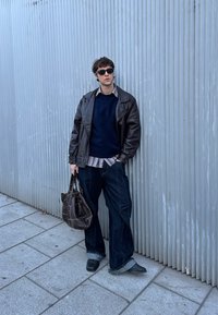 Giacca di pelle marrone, maglione lavorato blu, camicia a quadri, pantaloni in denim larghi, scarpe nere e una borsa marrone contro un muro ondulato.