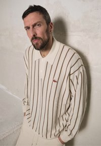 Suéter de malha de polo riscado em bege, com colarinho clássico, riscas verticais castanhas e um pequeno logótipo vermelho. O tecido tem uma textura suave.