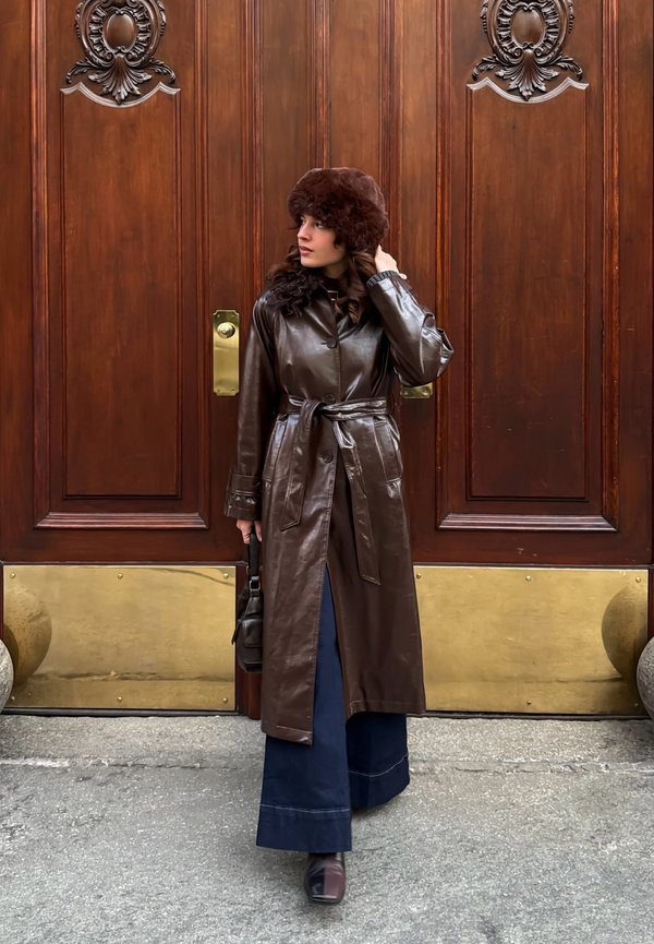 Vrouw in een bruin lederen trenchcoat, bontmuts en wijde broek staat voor grote houten deuren met gouden accenten, terwijl ze een handtas vasthoudt.