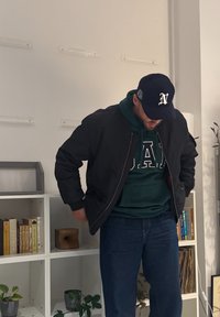 Jachetă bomber neagră peste o hanorac verde cu inscripții albe, blugi albaștri și o șapcă închisă la culoare, stând în fața unei rafturi.