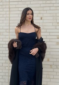 Femme portant une robe en dentelle bleu marine avec des bretelles fines et un manteau noir avec une bordure en fourrure foncée, debout devant un mur de briques claires.