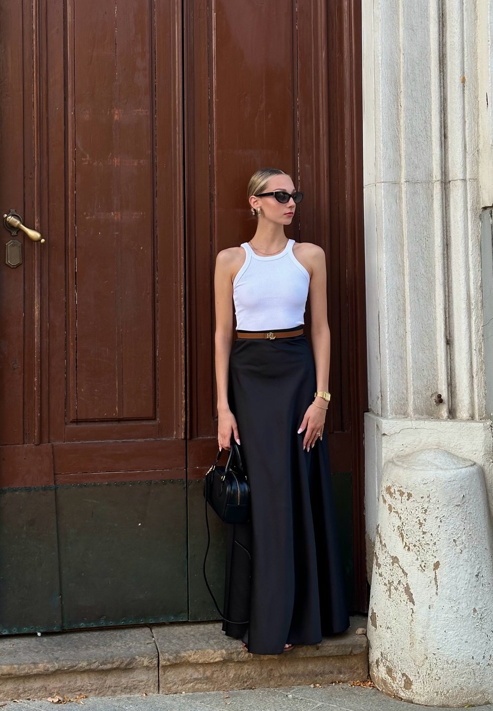 Haut sans manches blanc associé à une longue jupe noire, un sac à main noir et des lunettes de soleil, devant une porte en bois brun texturé.
