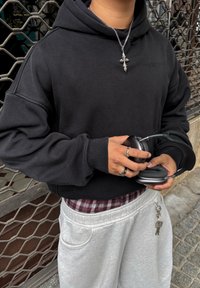 Personne portant un sweat à capuche noir et un collier croix argentée tient un casque audio noir tout en portant un pantalon gris clair avec un porte-clés.
