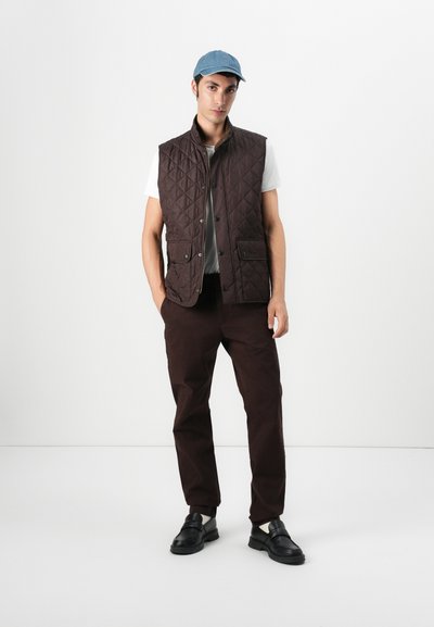 Chaleco acolchado marrón con un alto cuello, botones delanteros y dos bolsillos laterales, llevado sobre una camiseta blanca y combinado con pantalones marrones y zapatos negros.