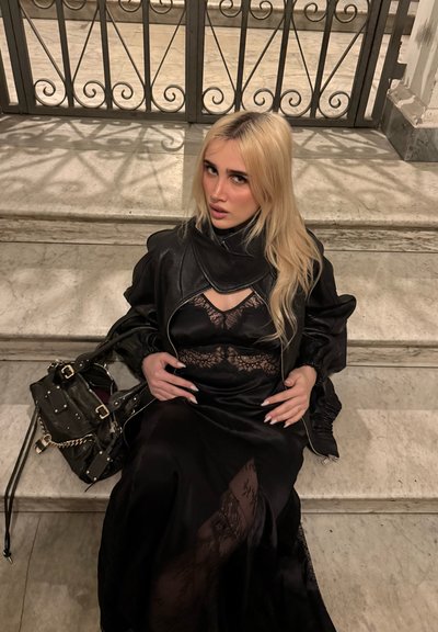 Mujer rubia con vestido de encaje negro y chaqueta de cuero sentada en escaleras de mármol, sosteniendo un bolso negro a su lado, con una barandilla ornamentada detrás.