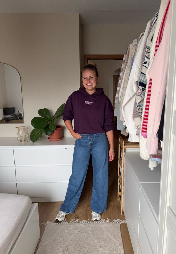 Femme souriante dans une chambre, portant un sweat à capuche violet, un jean bleu à jambes larges et des baskets, debout près de commodes blanches et de vêtements suspendus.