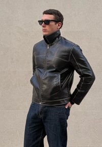 Άντρας που φοράει μαύρο δερμάτινο μπουφάν με ψηλό γιακά, σκούρα τζιν και γυαλιά ηλίου, στέκεται μπροστά σε μπεζ πλακιδωτό τοίχο με τα χέρια στις τσέπες.