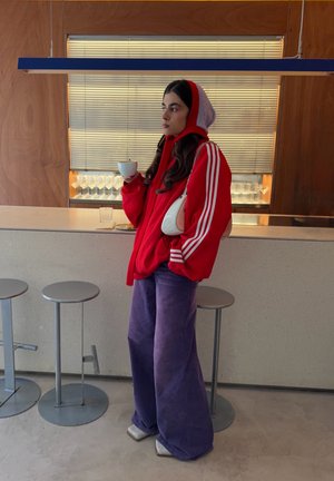 Persona con sudadera roja con capucha y pantalones anchos morados sostiene una taza blanca, de pie junto a un mostrador de bar con taburetes y cristalería al fondo.